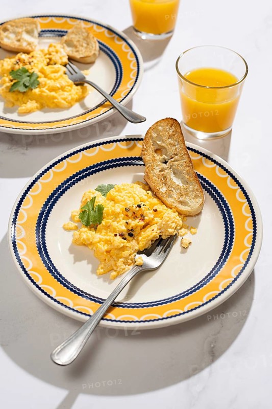 A vibrant breakfast scene featuring creamy scrambled eggs garnished with herbs, served on a colorful plate. Accompanied by toasted bread and a refreshing glass of orange juice