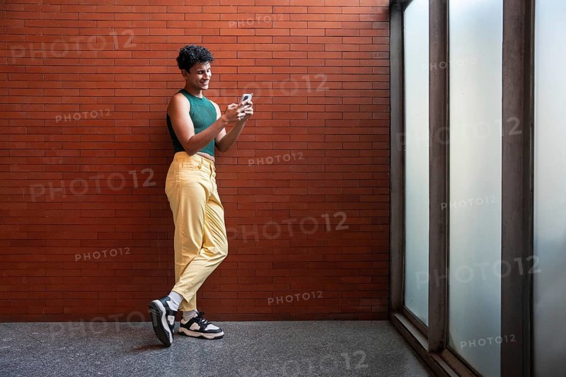 A person stands casually, texting on their phone against a red brick wall, wearing a green top and yellow pants, conveying a relaxed, modern vibe
