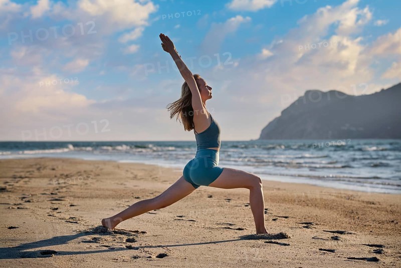A woman is performing Virabhadrasana 1, or Warrior 1 pose, on the sandy beach as the sun sets. The calm sea and distant hills create a tranquil backdrop for her yoga practice