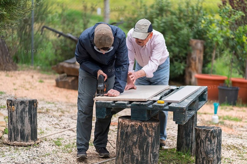 Senior father and son working together using a jigsaw on a woodworking project in an outdoor setting, demonstrating a meaningful bonding activity through carpentry