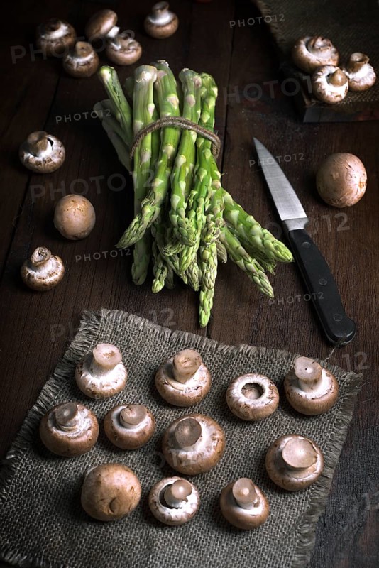 Fresh asparagus bundled with twine and mushrooms artfully arranged on a dark wooden table with burlap. A knife is placed beside, creating a rustic kitchen scene and vegetables