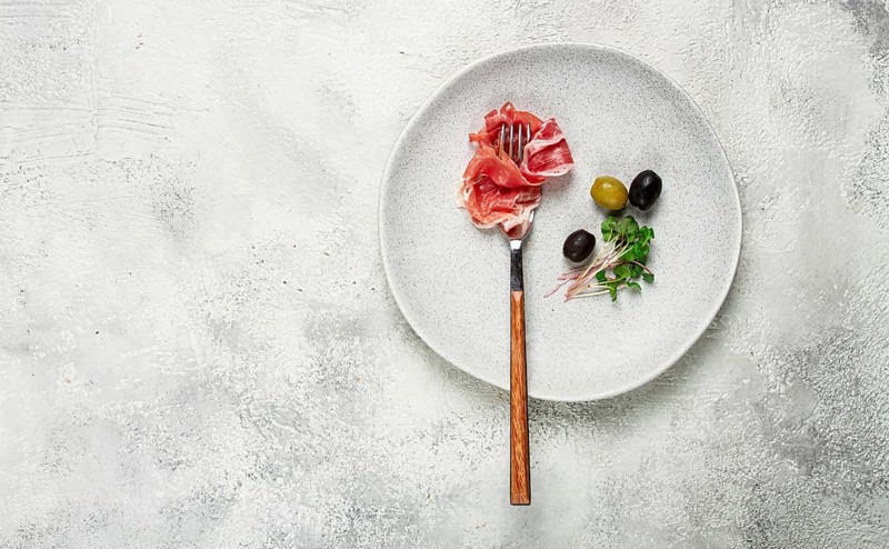 Prosciutto on a fork, top view, olives and micro-greens on a plate, light background, no people