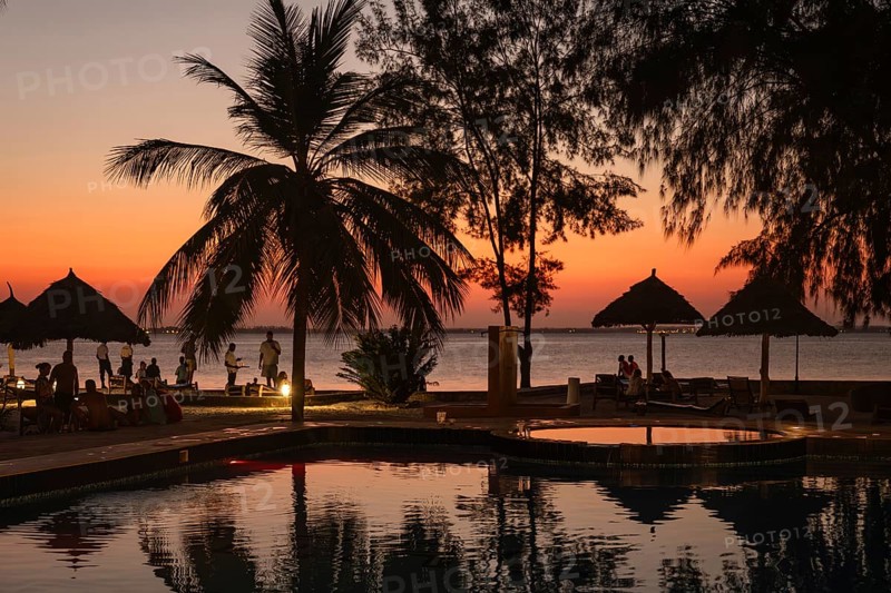 MICHAMVI KAE, ZANZIBAR - OCTOBER 19, 2025: Silhouetted palm trees and thatched umbrellas frame a luxury pool at sunset, with tourists relaxing by the ocean in Zanzibar