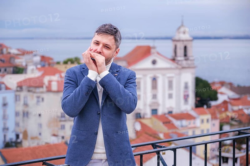 Man musician in a blue blazer and white sweater playing blues on a harmonica with eyes closed, standing outdoors in Lisbon city street, Portugal