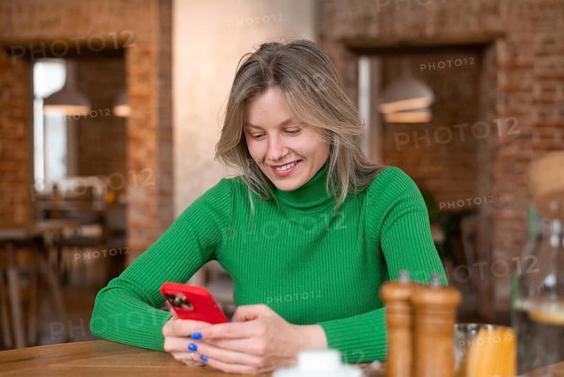 A woman in a green sweater smiles while using her phone at a cozy cafe. In the foreground, table items suggest a relaxed breakfast atmosphere