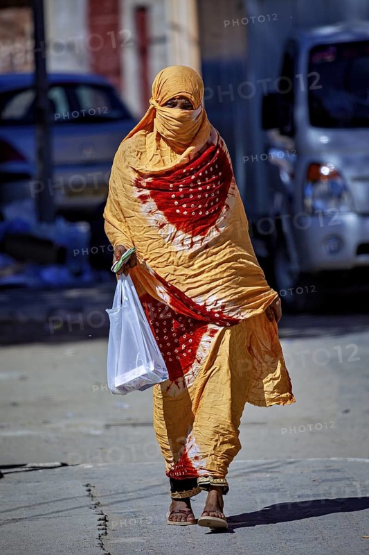A woman in a yellow and orange traditional dress carries a shopping bag, Veiled woman at the Tamanrasett festival in the Sahara in Algeria