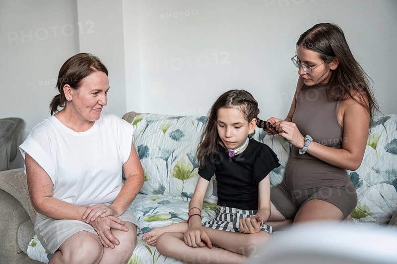A warm family scene with a disabled girl with a tracheostomy and her caretakers. One is braiding her hair while another shares a comforting moment, highlighting love and support