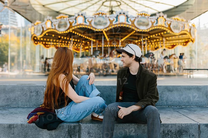 A young couple relaxes on concrete steps near a brightly lit carousel in Brooklyn Bridge Park. The colorful, lively background creates a cheerful atmosphere for their casual interaction in New York