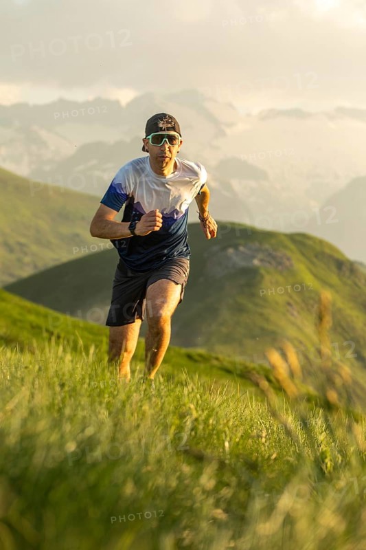 A determined man with sunglasses and a hat runs energetically through a lush, mountainous trail, embodying the spirit of adventure and athleticism on a sunny day