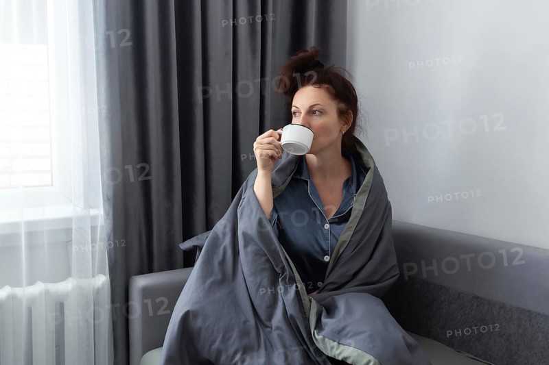 A woman in pajamas, wrapped in a gray blanket, enjoys her morning coffee on a sofa. Soft window light brightens the room, creating a peaceful and relaxed atmosphere
