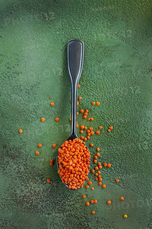 Bright orange lentils fill a silver spoon, with some lentils scattered on a green textured surface, showcasing the vibrant color and texture of these legumes