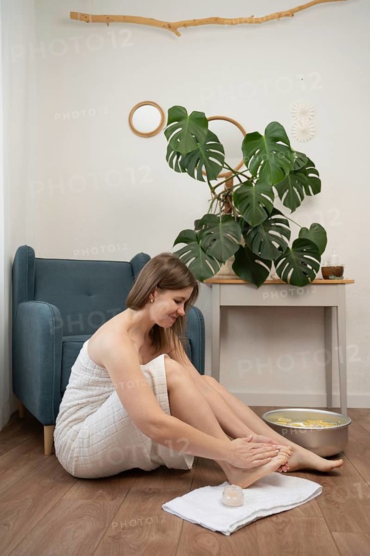 A woman in a towel enjoys an at-home spa session, applying skincare after a relaxing citrus foot soak. The serene setting features minimalist decor, a monstera plant, and calming natural elements
