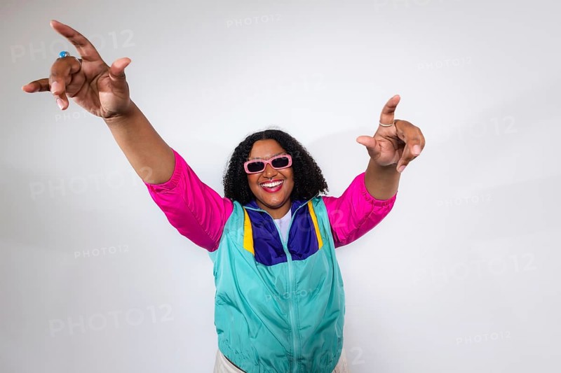 A black woman in a colorful 80s-inspired outfit, wearing pink sunglasses, exudes happiness and excitement. The vibrant colors and dynamic pose capture a joyful retro vibe