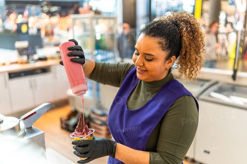 Smiling woman employee working in a food service business, wearing gloves and an apron, adding a sweet topping to a chocolate and vanilla ice cream in a cup