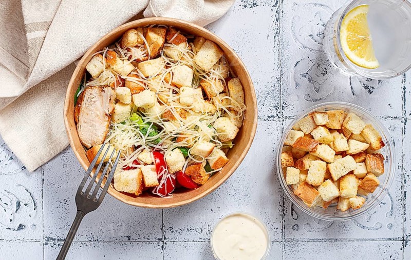 Caesar salad in a paper container, take it on the way, with sauce and croutons, on the table, snack, natural light, no people