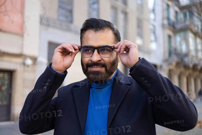 Indian man in a professional outfit adjusts his eyeglasses while standing on a city street. He's looking away with a confident expression