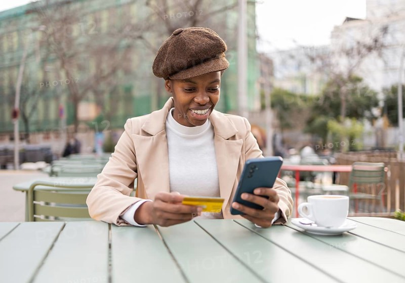 A stylish black woman in an urban setting, enjoying coffee while shopping online with her phone and credit card The vibrant city ambiance enhances her modern lifestyle