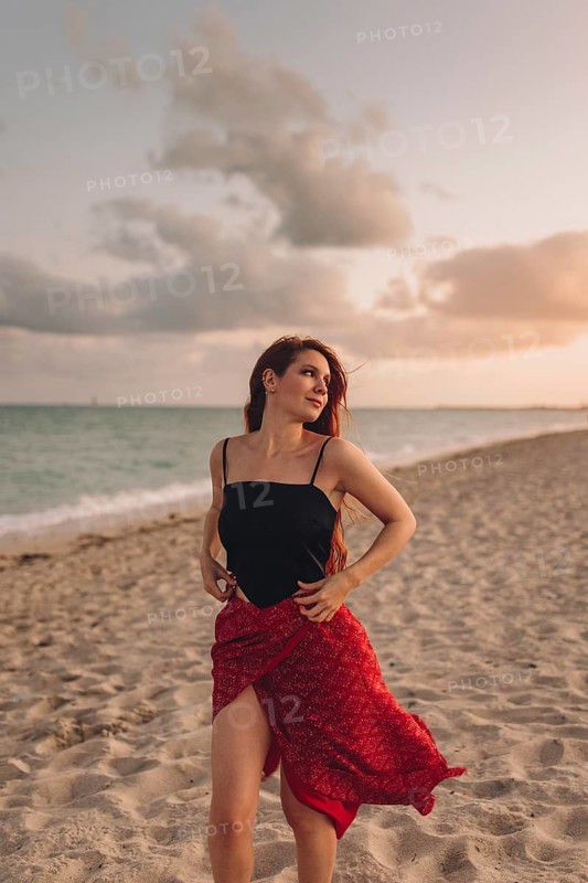 A young woman with red hair stands gracefully on a sandy beach in Miami, dressed in a stylish black top and a flowing red skirt. The gentle breeze and serene background of clouds and sea create a tranquil atmosphere during her beach layover