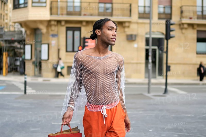 Stylish man in a mesh top confidently walks through a city street. He embodies LGBTQIA+ pride, showcasing vibrant fashion and self-expression while holding a shopping bag