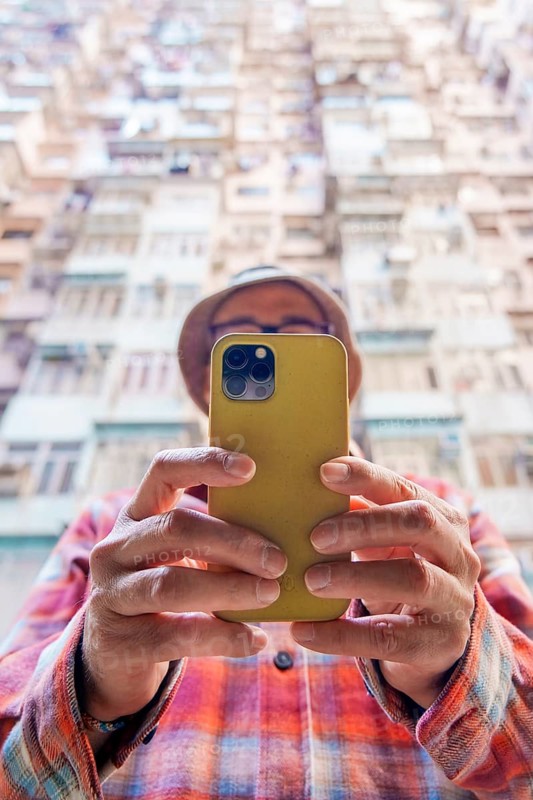 From below an unrecognizable man in a hat uses a smartphone in front of a dense Hong Kong residential backdrop