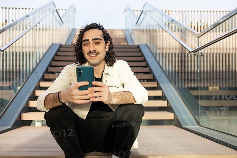 Relaxed man with a mustache sits on outdoor steps, engaging with a smartphone Dressed casually, exuding a modern urban vibe, surrounded by contemporary architecture