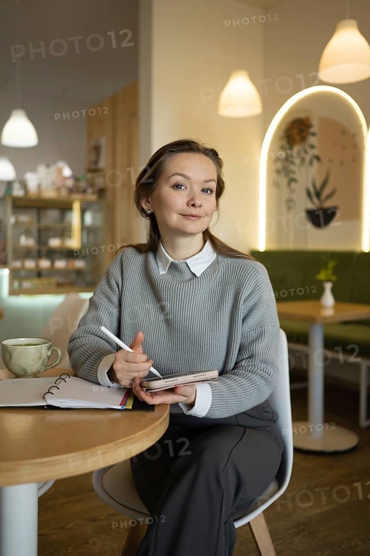 A woman sits in a cozy cafe, holding a tablet pencil and smiling, surrounded by modern decor. A notebook and coffee cup are on the table, creating a relaxed ambiance