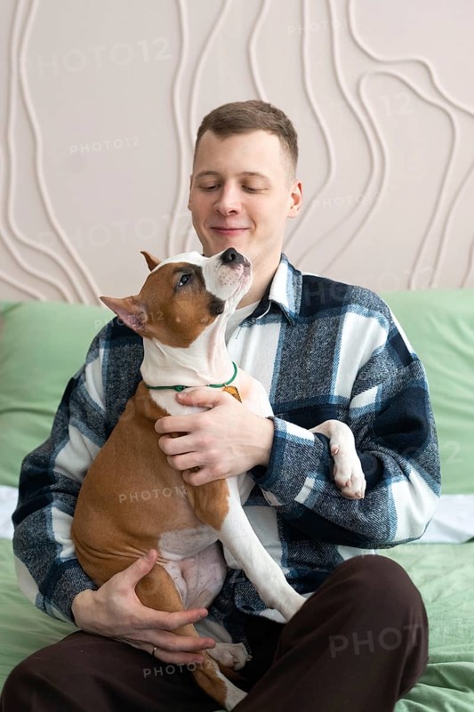 A young man relaxes at home with a playful brown and white dog in his lap, enjoying a moment of affection and companionship