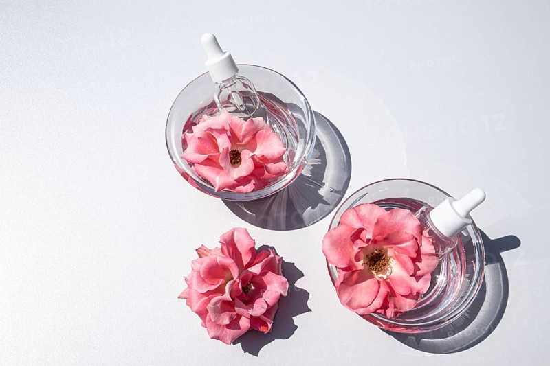 Top view of two glass bowls filled with clear rose water, each containing a pink rose and a dropper bottle serum, on a white background, creating a minimalist aesthetic