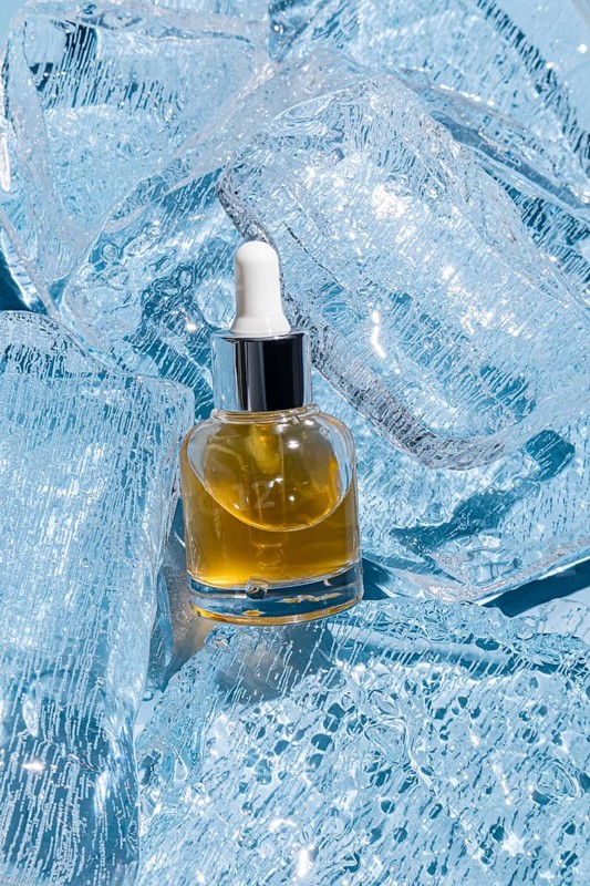 An elegant glass serum bottle with a dropper filled with golden liquid, embedded in a bed of sparkling ice cubes. This image captures the essence of luxury skincare kept chilled