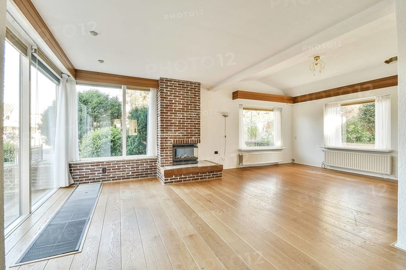 A spacious living room with large windows allowing for natural light, featuring wooden floors and a cozy brick fireplace, ideal for relaxation