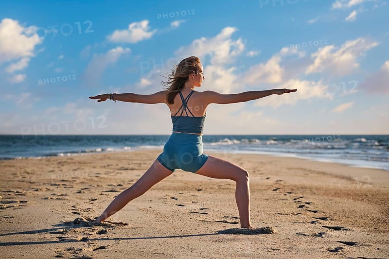 A woman performs the Warrior 2 Virabhadrasana 2 pose on the beach, enjoying the calm sunset and ocean breeze during her yoga practice