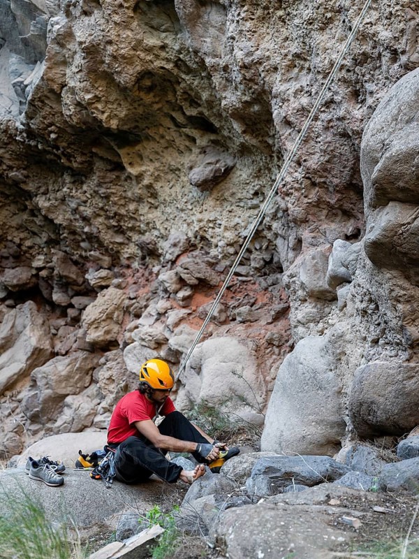 A climber prepares for a traditional climb on a rocky natural cliff. Wearing safety gear, they focus on lacing up climbing shoes, surrounded by rugged terrain and rock formations