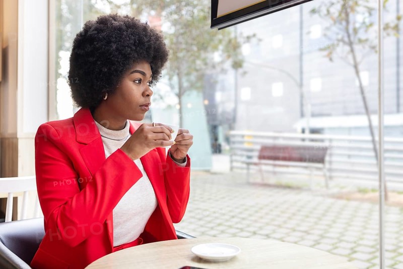 A woman in a vibrant red jacket enjoys a cup of coffee, seated in a modern cafe. The outdoors visible through large windows add to the serene atmosphere