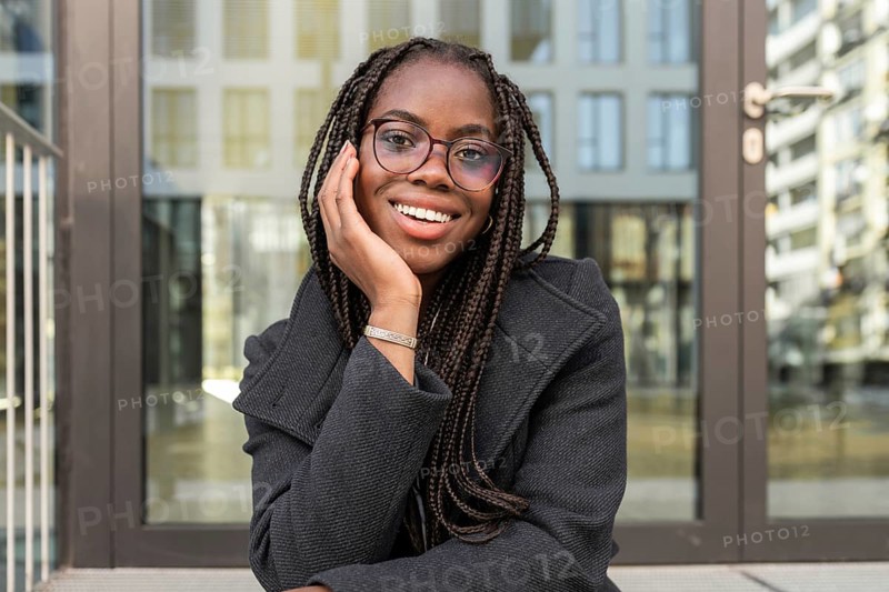 Smiling young African American businesswoman with braids, dressed in a professional suit, taking a break outdoors while seated comfortably