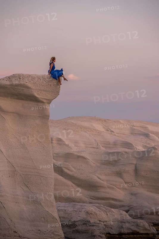 Unrecognizable woman in a flowing blue dress perched on a white rock formation at Sarakiniko, island of Milos, Greece, during sunset