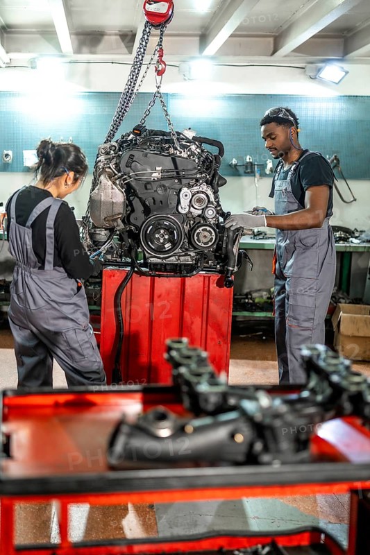 Two auto mechanics inspecting a car engine hanging from a chain hoist in a professional workshop