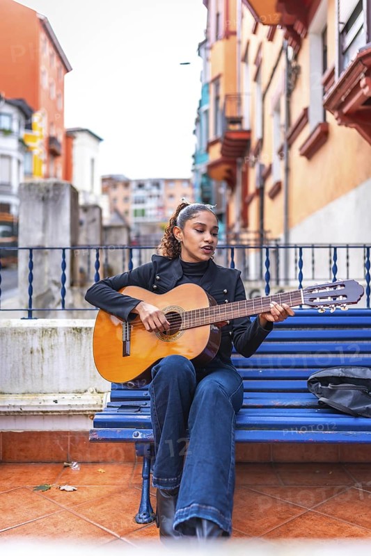 Vertical portrait of a beauty woman african american musician playing guitar sitting on a city bench