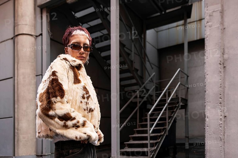 A stylish woman wearing a faux fur coat and sunglasses stands confidently in an urban alley. The setting features industrial stairs and a modern concrete backdrop