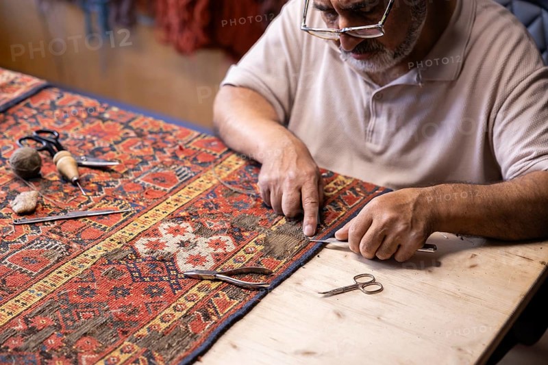 An Iranian carpet-restoration expert works meticulously in his Salzburg workshop, using traditional tools to repair an intricate, colorful carpet