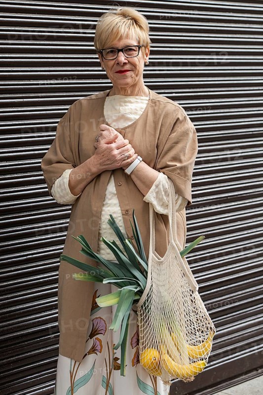 Fashionable woman stands confidently holding a mesh grocery bag with fresh produce. She wears glasses, a beige outfit, and a floral skirt, showcasing style and sustainability