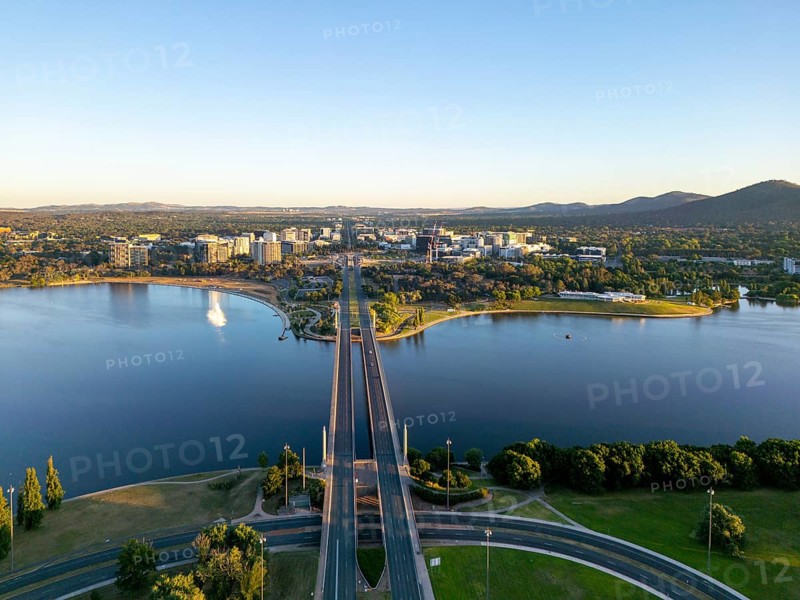 From above, a drone view of Canberra's cityscape, highlighted by meandering roads and the large Molonglo River. The scene is complemented by distant hills and neatly organized urban areas, providing a serene yet dynamic urban panorama