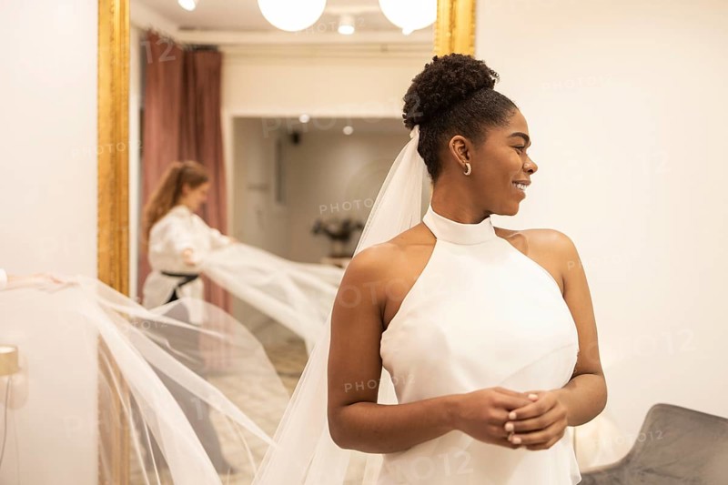 A bride smiles elegantly as she tries on a sophisticated wedding dress in a warmly lit bridal shop. An attendant adjusts her veil, ensuring every detail is perfect