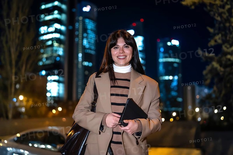 Businesswoman posing with a tablet, smiling brightly for the camera in front of a blurry background of illuminated skyscrapers and city lights, symbolizing career success and urban professional life