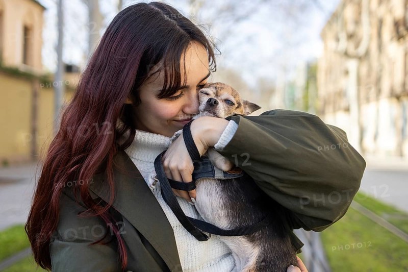 A woman warmly cuddles her small chihuahua outdoors, showcasing a tender moment of affection. The setting hints at a pleasant day, reflecting joy and companionship