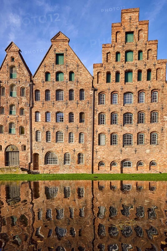 Historic salt storehouse, brick building with windows and doors against blue sky with veil clouds, reflection on the water surface of the river Trave, Hanseatic city of Lübeck, Schleswig-Holstein, Germany