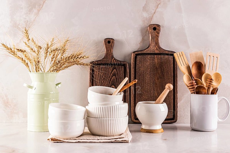 A collection of rustic wooden kitchen utensils, cutting boards, and white ceramic bowls arranged neatly with a vase of wheat, set against a light marble backdrop