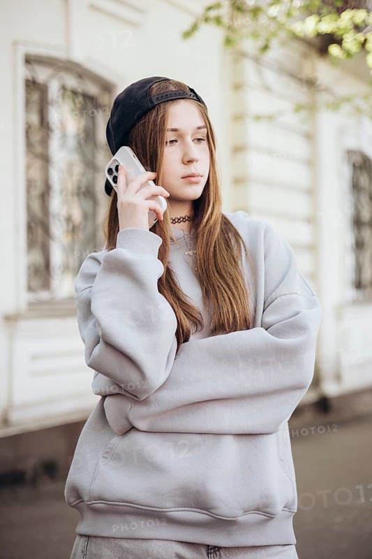 Teenage girl wearing a casual outfit and a cap, holding a smartphone to her ear, standing outside on a sunny day, appears focused and engaged in conversation