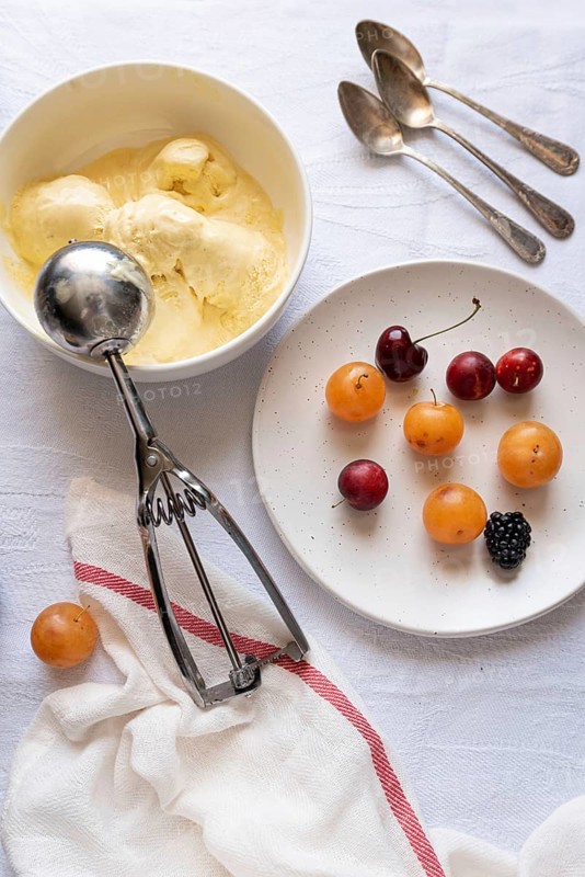 Top view of a refreshing dessert layout with a bowl of creamy vanilla ice cream, a plate of vibrant cherries, apricots, and a blackberry laid on a white textured tablecloth alongside vintage spoons