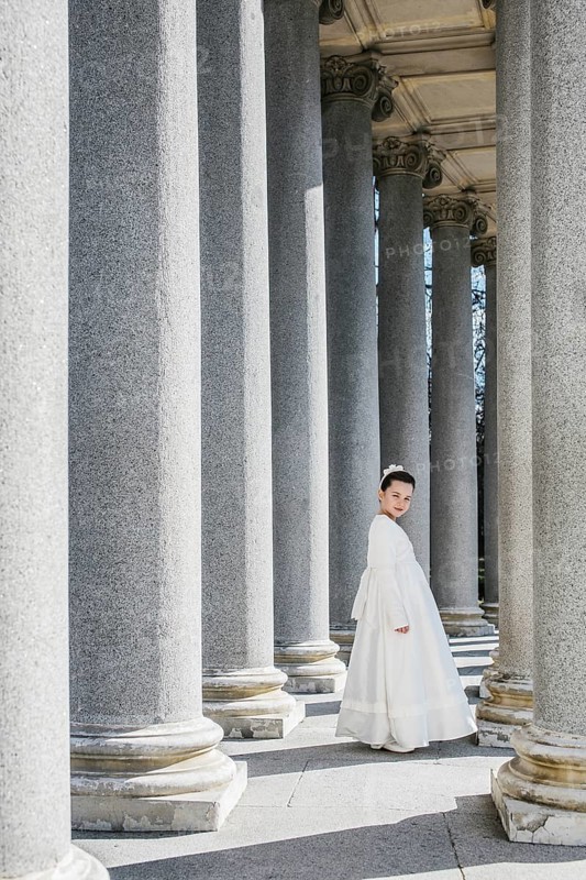 A young girl stands gracefully in a First Communion Day dress, surrounded by towering stone columns. The serene setting highlights the beauty of the traditional attire