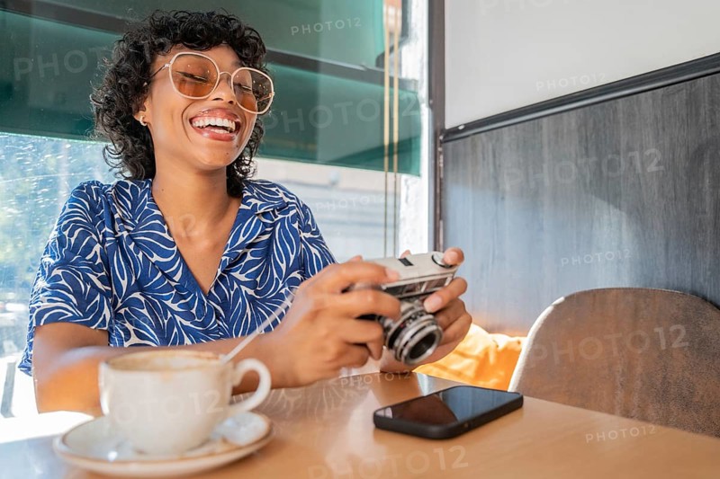 A cheerful woman in a blue patterned shirt enjoys coffee in a cafe. She holds a vintage camera, her smartphone nearby. Sunlight streams in, creating a relaxed atmosphere